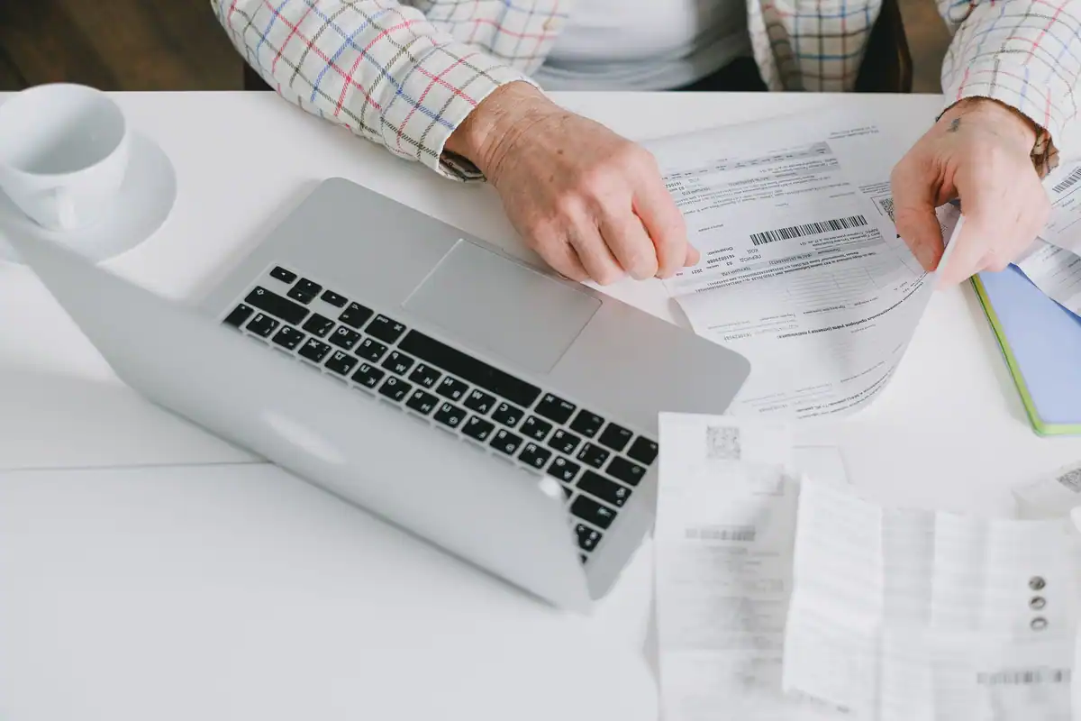 Mâini care răsfoiesc un fluturaș de salariu lângă un laptop, pe un birou alb