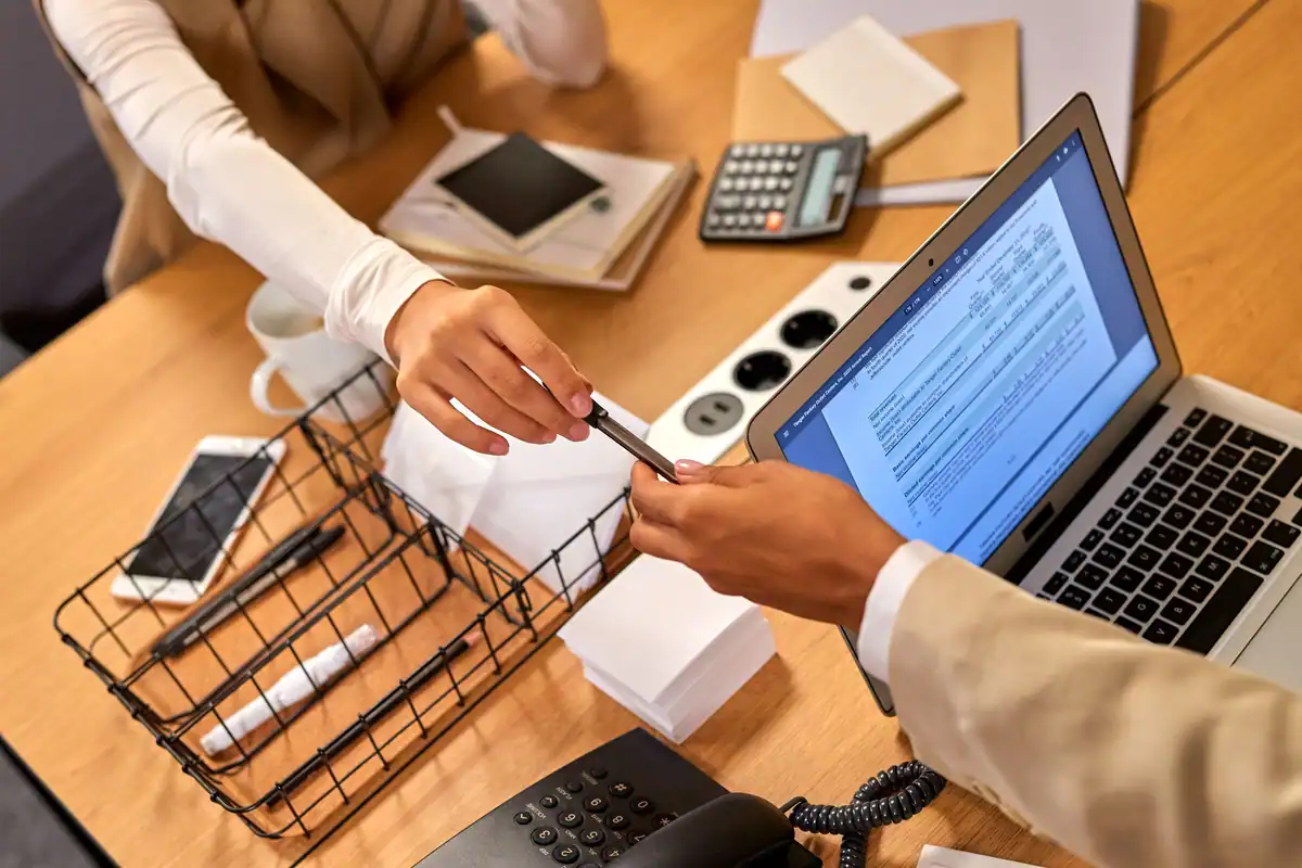 Doi oameni la un birou, unul predă un document celuilalt, lângă un laptop și un calculator