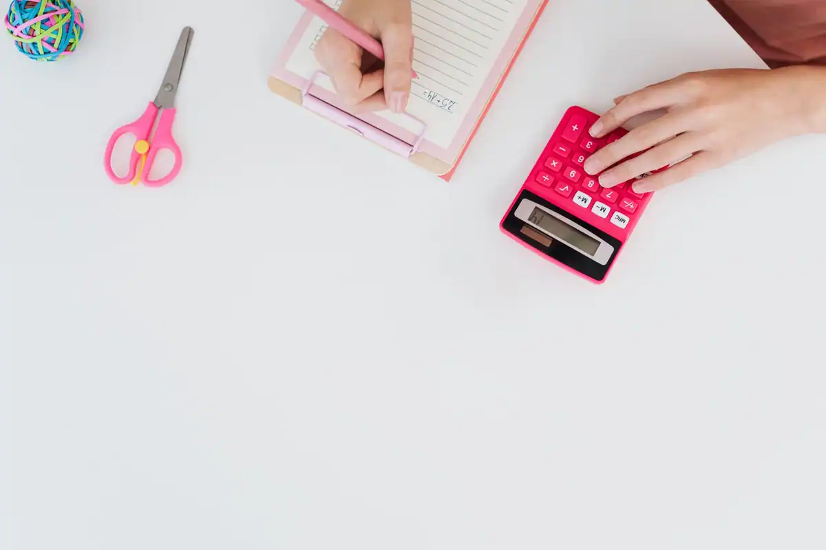 Mână care calculează cu un calculator roz, pe un birou alb, lângă foarfecă și caiet