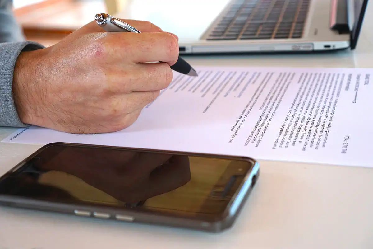 Mână care semnează un contract, lângă un telefon și un laptop, pentru veniturile din activități